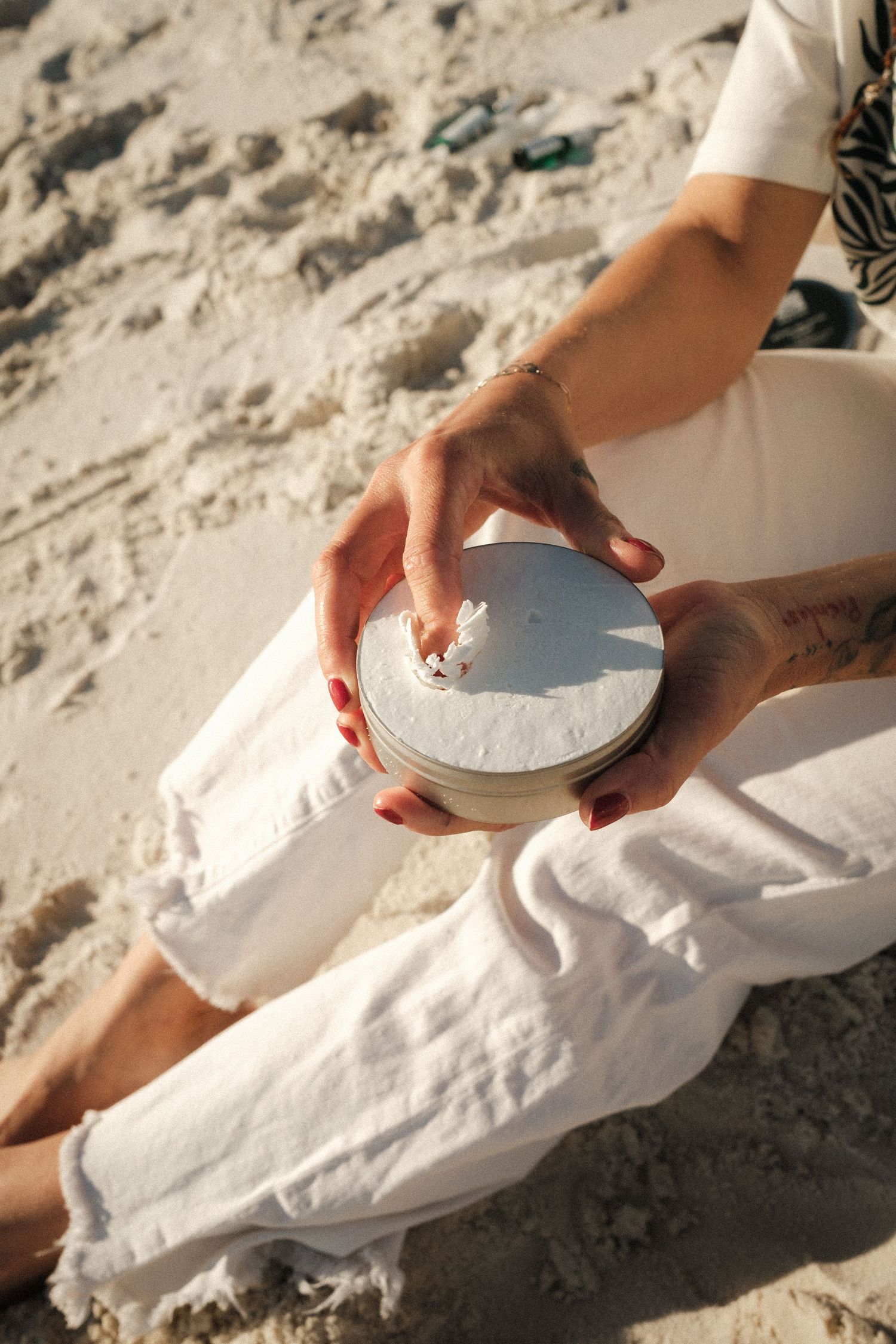 Woman with red nails and tattoo holds a white round candle tin while sitting on sandy beach in white linen outfit.