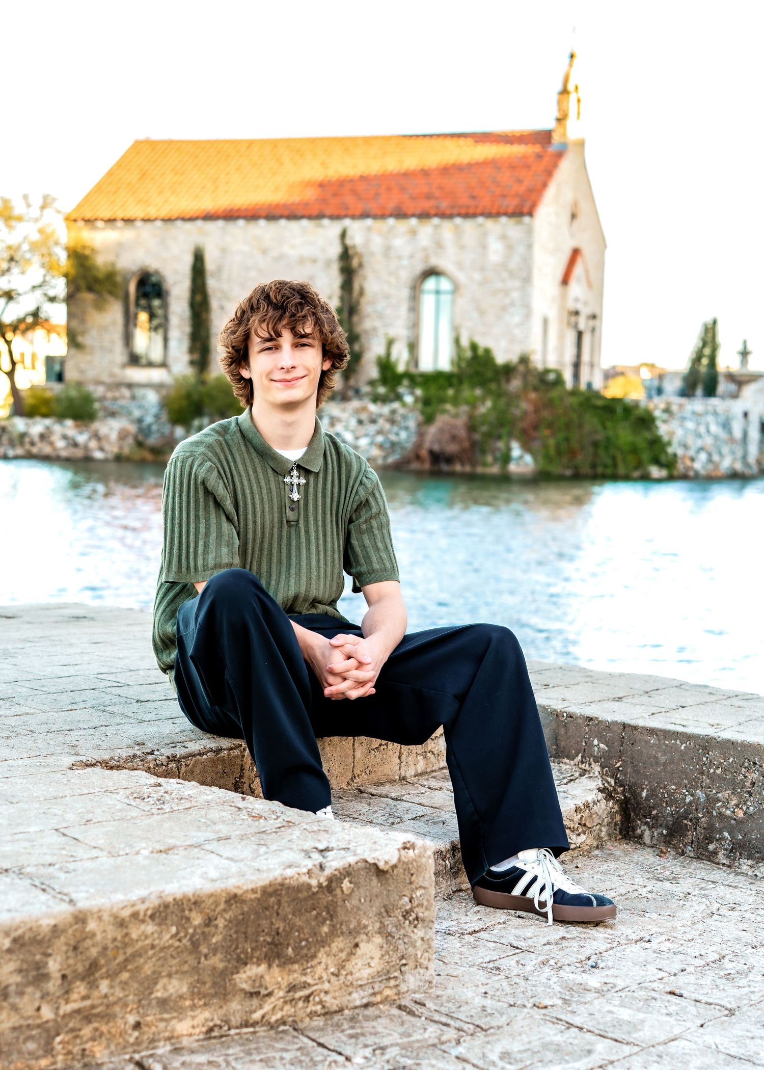 Young man with curly hair in green shirt stands on metal staircase against stone wall.