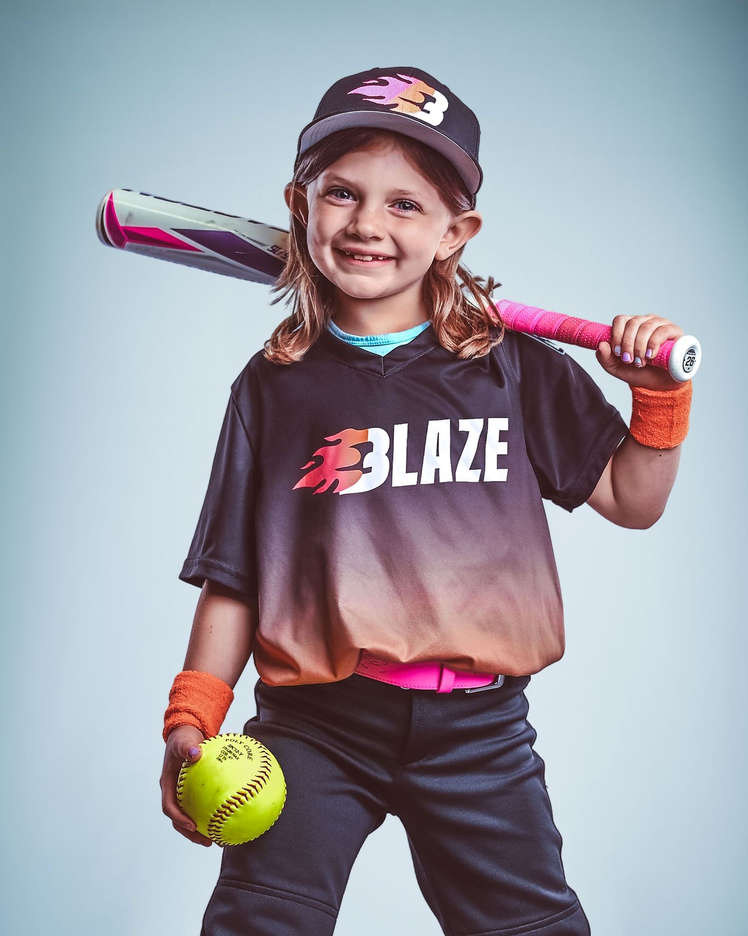 Young girl in Blaze softball uniform smiling, holding yellow ball and pink bat over shoulder, wearing team cap.