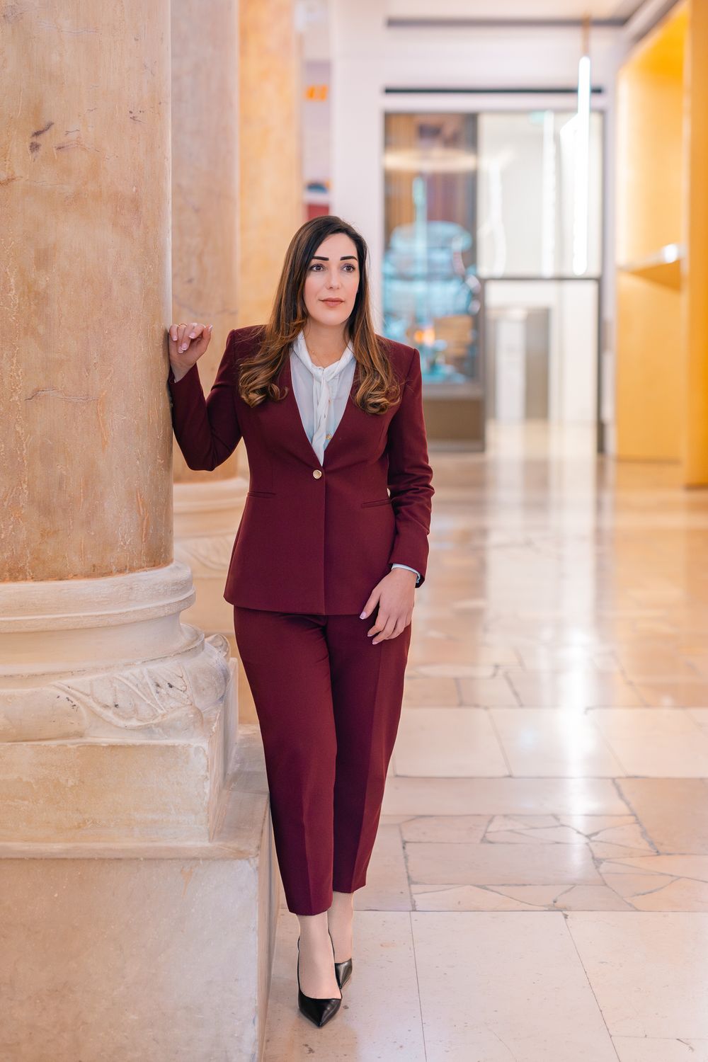 Professional woman in burgundy suit leaning against marble column in elegant office hallway.