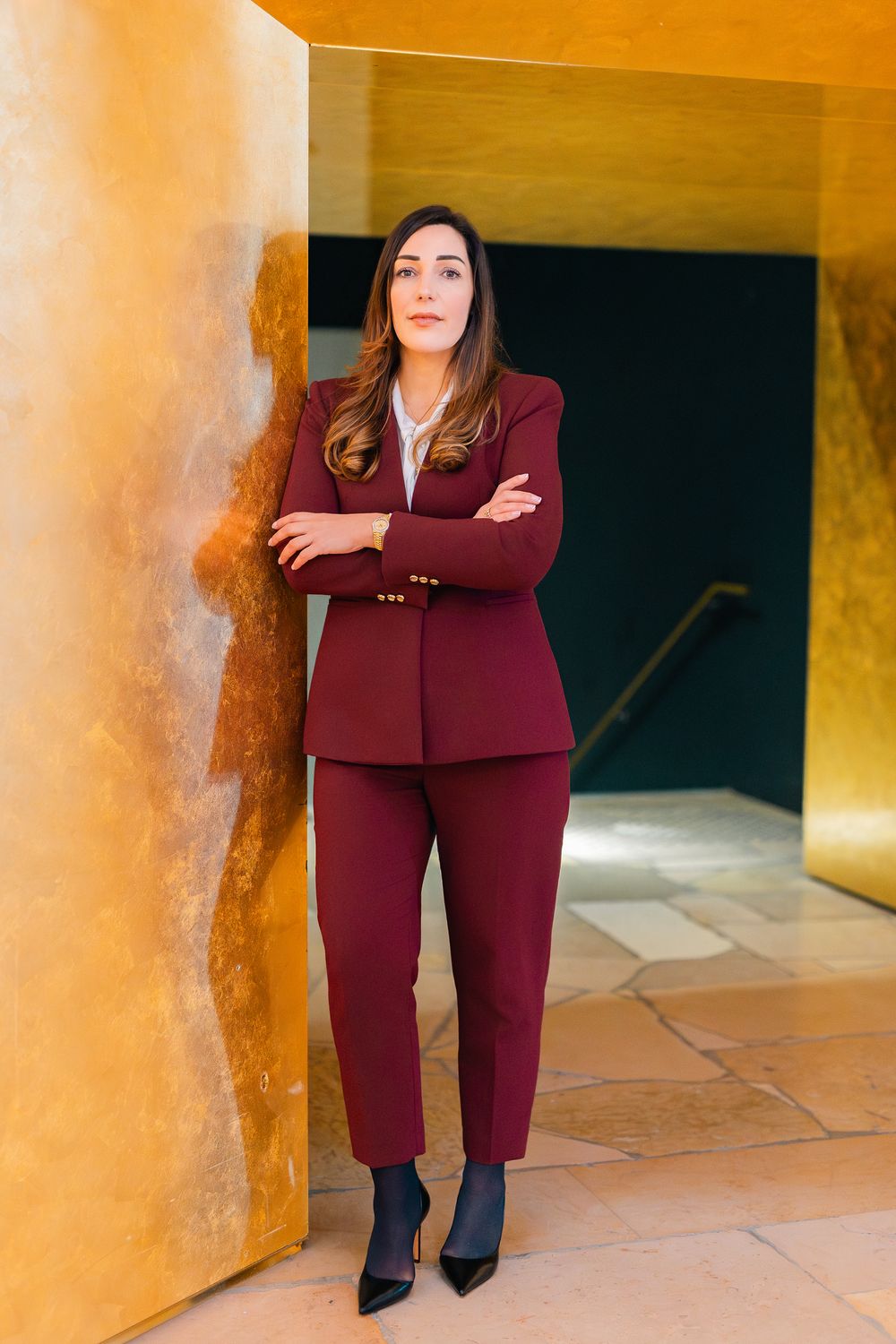 Woman in burgundy pantsuit stands confidently with arms crossed leaning against golden wall in modern architectural space.