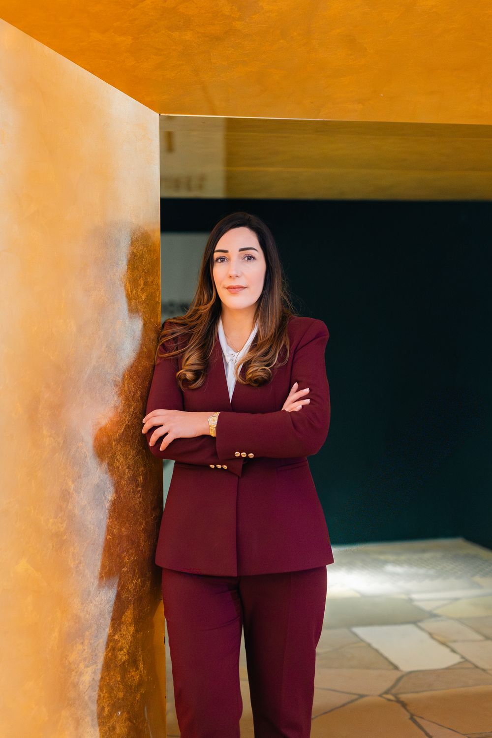 Professional woman wearing maroon suit poses with crossed arms against rustic golden wall in contemporary setting.