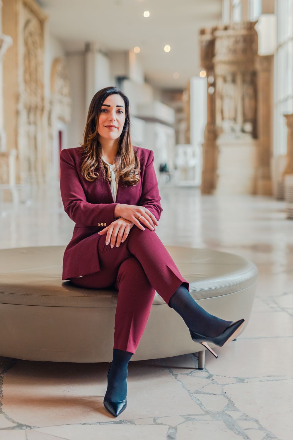 Woman in burgundy suit sitting confidently on a round ottoman in an elegant marble hall.
