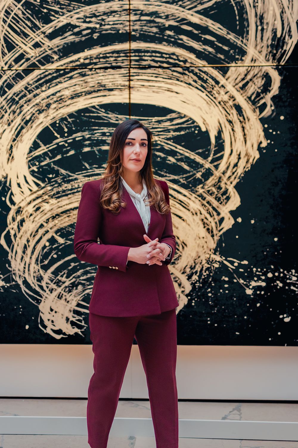 Businesswoman in burgundy suit stands before dramatic black and gold abstract artwork in elegant interior space.