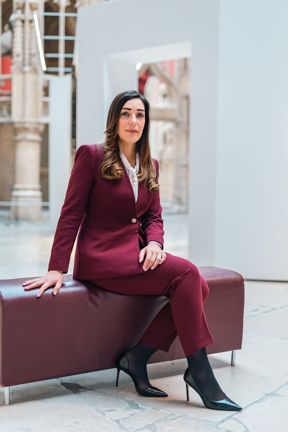 A professional woman in a burgundy suit sits confidently on a leather bench in a modern gallery space.