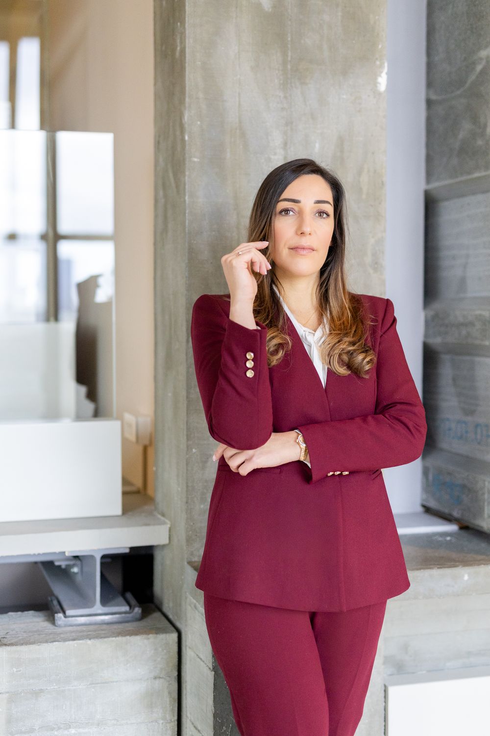 A professional woman in a burgundy suit leans against a concrete pillar, looking thoughtfully to the side.