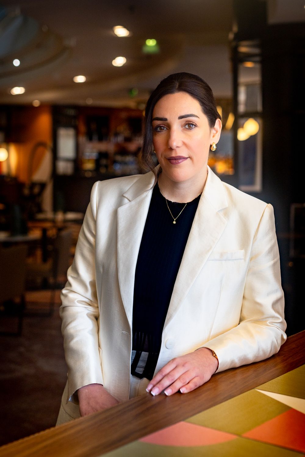 Professional woman in white blazer leaning on a bar counter in an elegant restaurant interior.