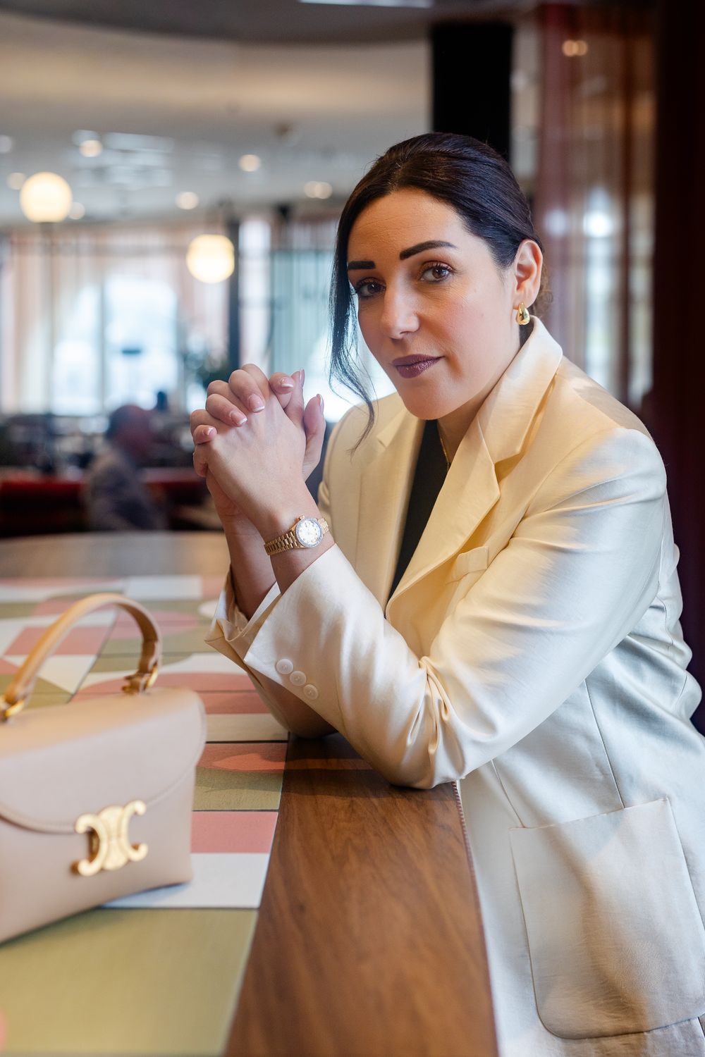 Elegant woman in cream blazer poses at wooden table with Celine handbag in upscale hotel lobby.