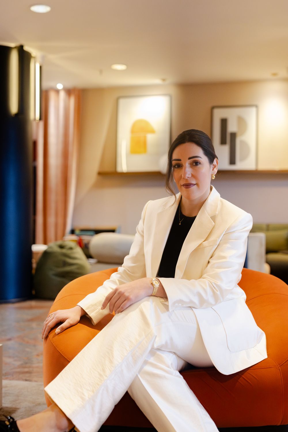 Confident businesswoman in white suit sitting on an orange ottoman in a modern hotel lobby.
