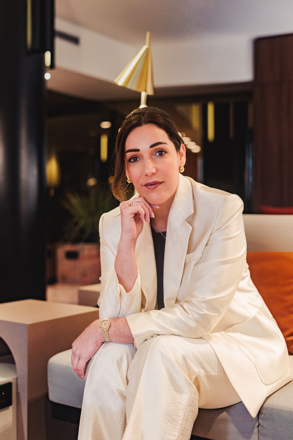 Elegant woman in white suit sitting confidently in modern interior with gold lamp.
