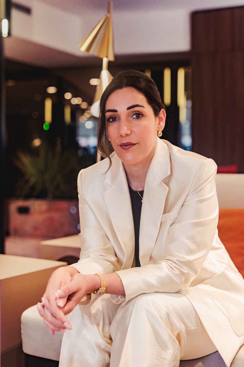 Stylish woman in cream blazer with gold jewelry sitting in a warmly lit contemporary lounge.