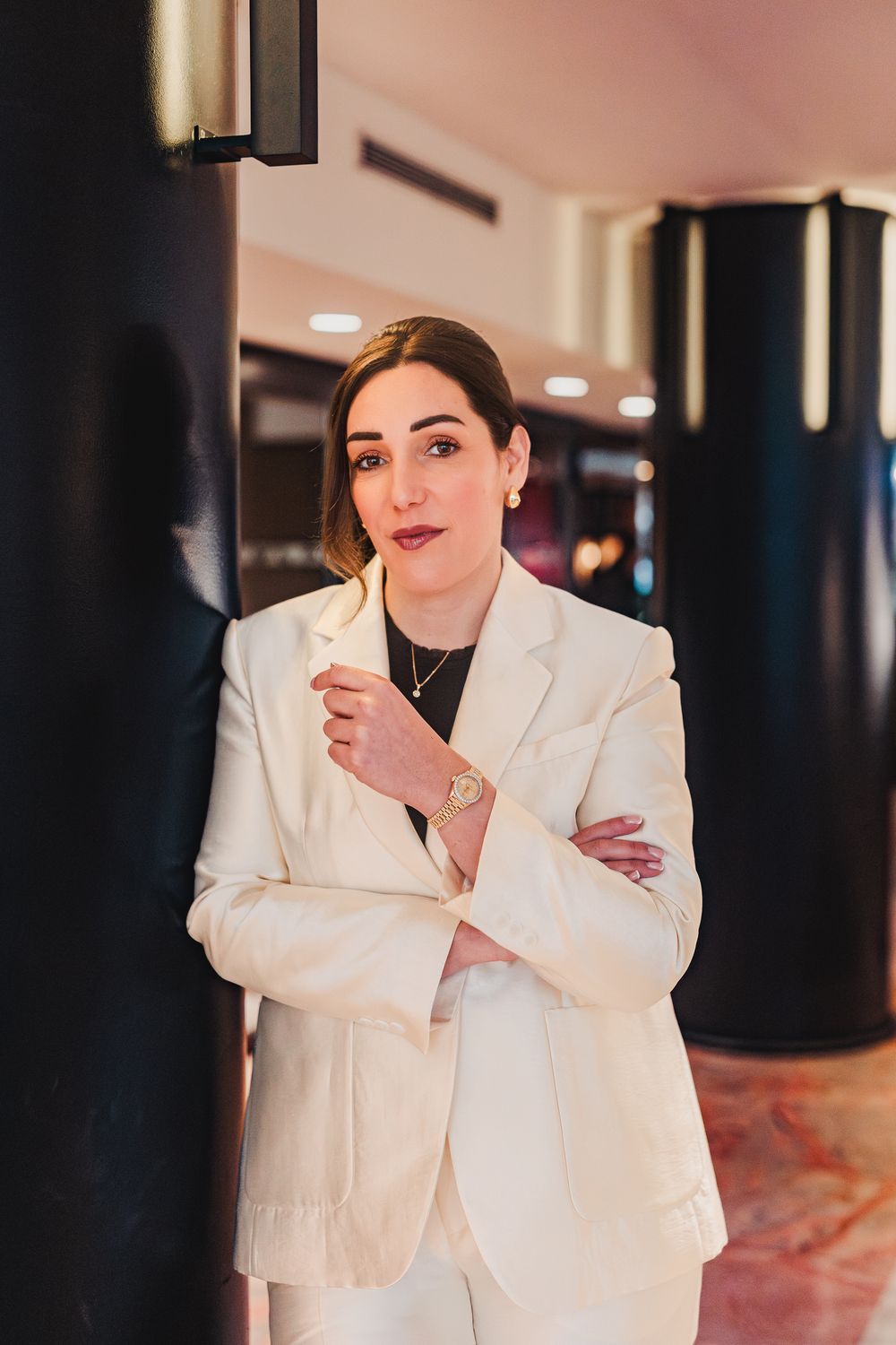 Elegant woman in white suit leaning against a dark column in a sophisticated venue interior.