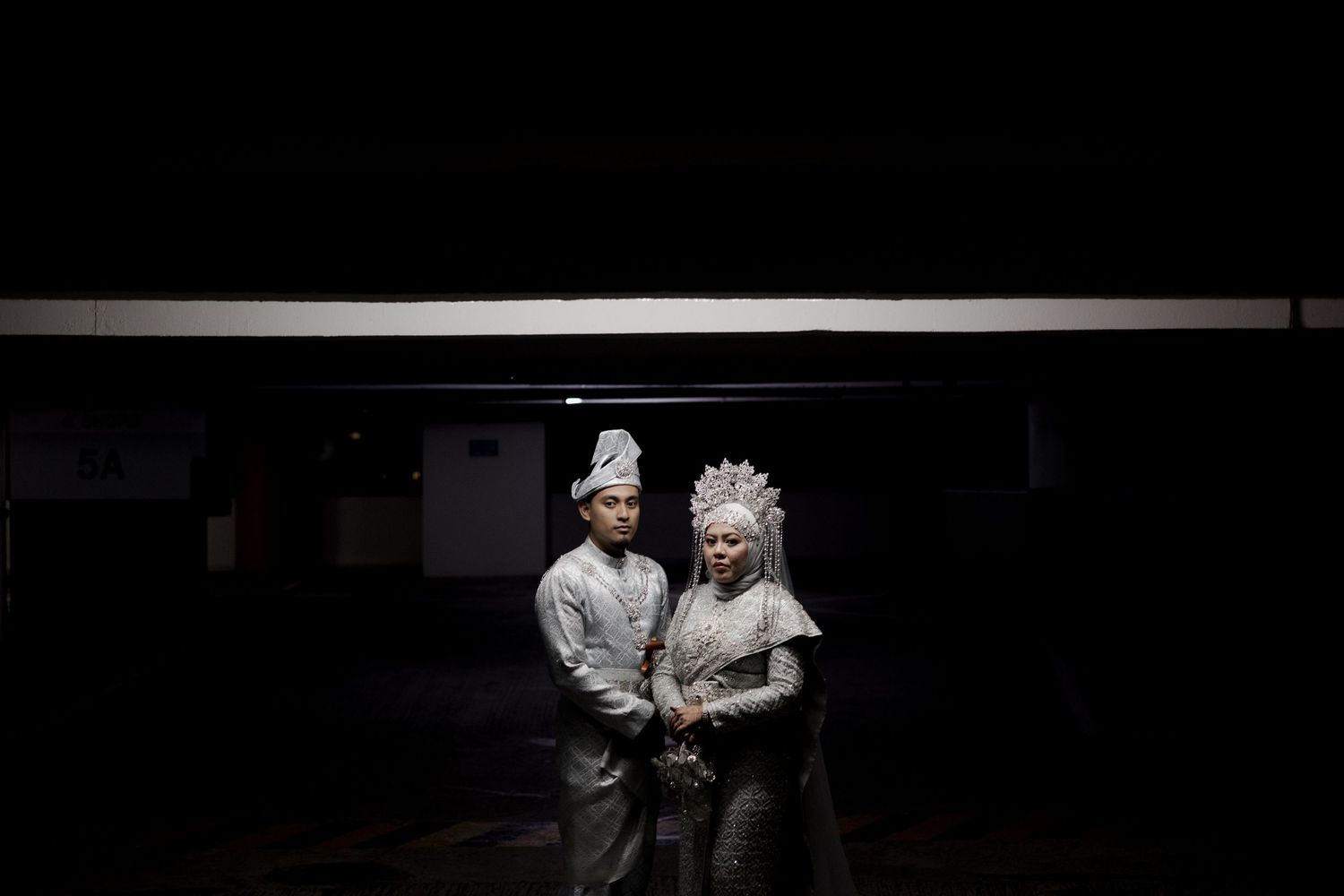 Bride in white lace gown stands beside groom in black tuxedo seated on red chair against metallic wall.
