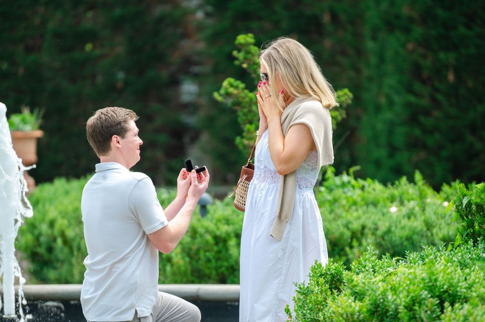 Man kneeling on one knee proposing to a surprised woman outdoors, holding a ring box in a lush garden setting.