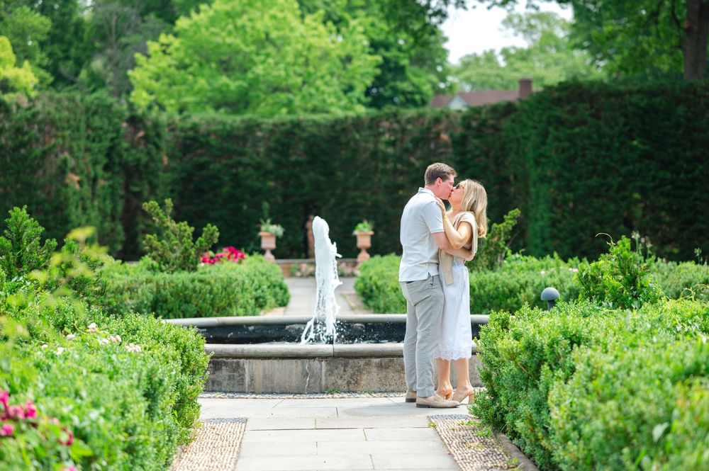 Couple sharing a romantic kiss by a garden fountain surrounded by lush green hedges and colorful flowers.