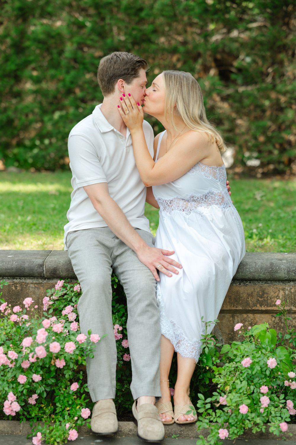 Couple sharing a romantic kiss while sitting on a stone wall surrounded by pink roses in a lush garden.