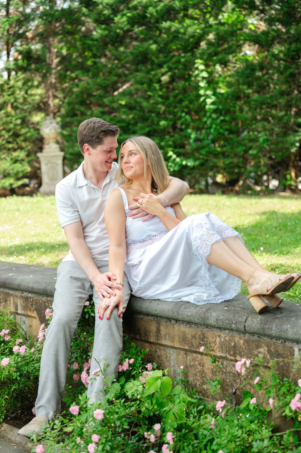 Couple sitting on a stone garden wall surrounded by pink roses and lush greenery, smiling at each other.