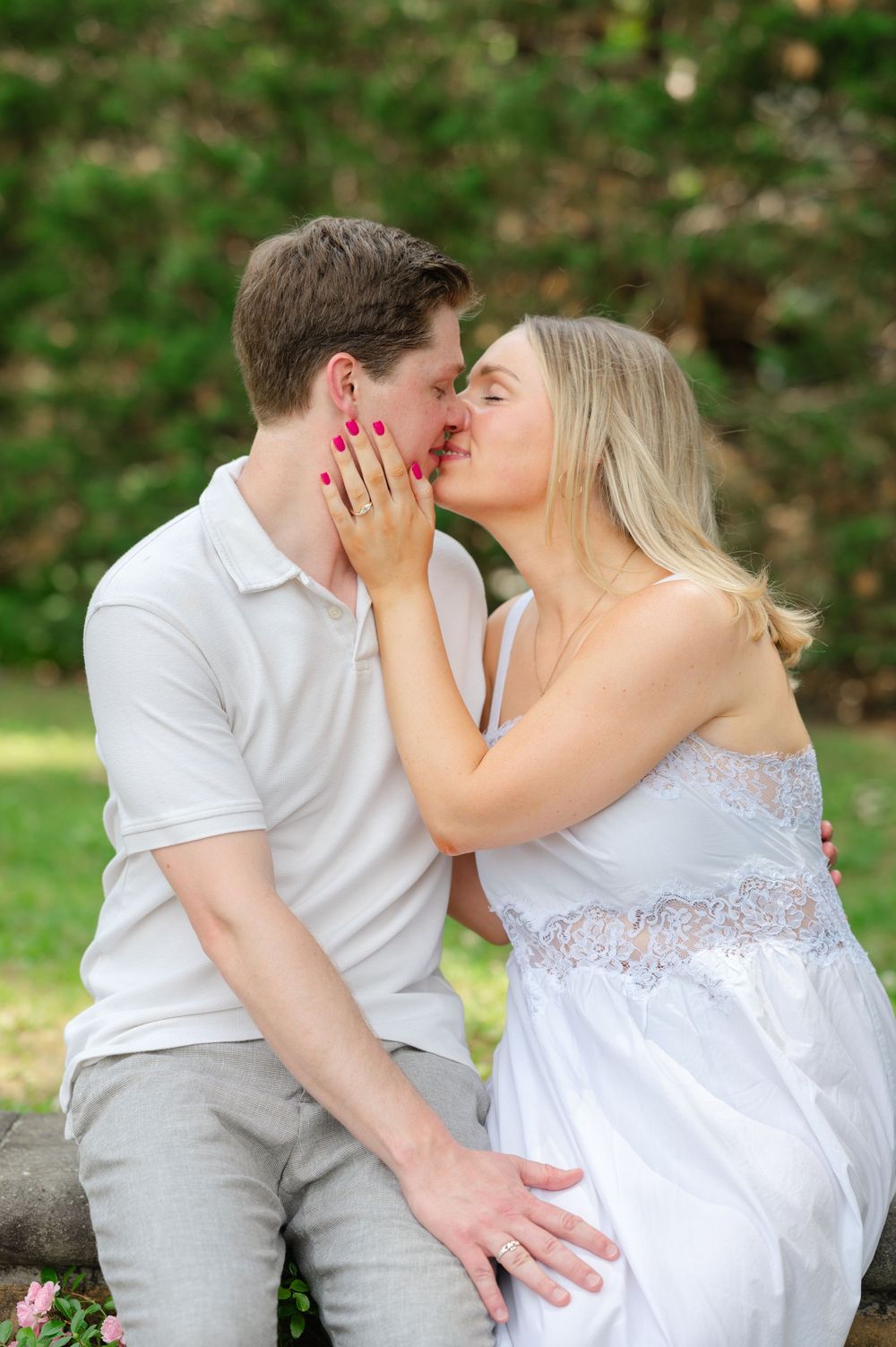 Couple sharing a romantic kiss outdoors in a lush green park, woman wearing white dress showing engagement ring.