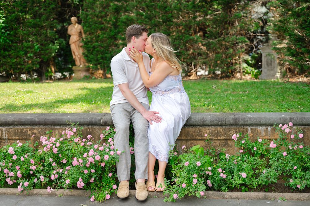 Couple sharing a romantic kiss on a stone garden bench surrounded by pink flowers and lush greenery.