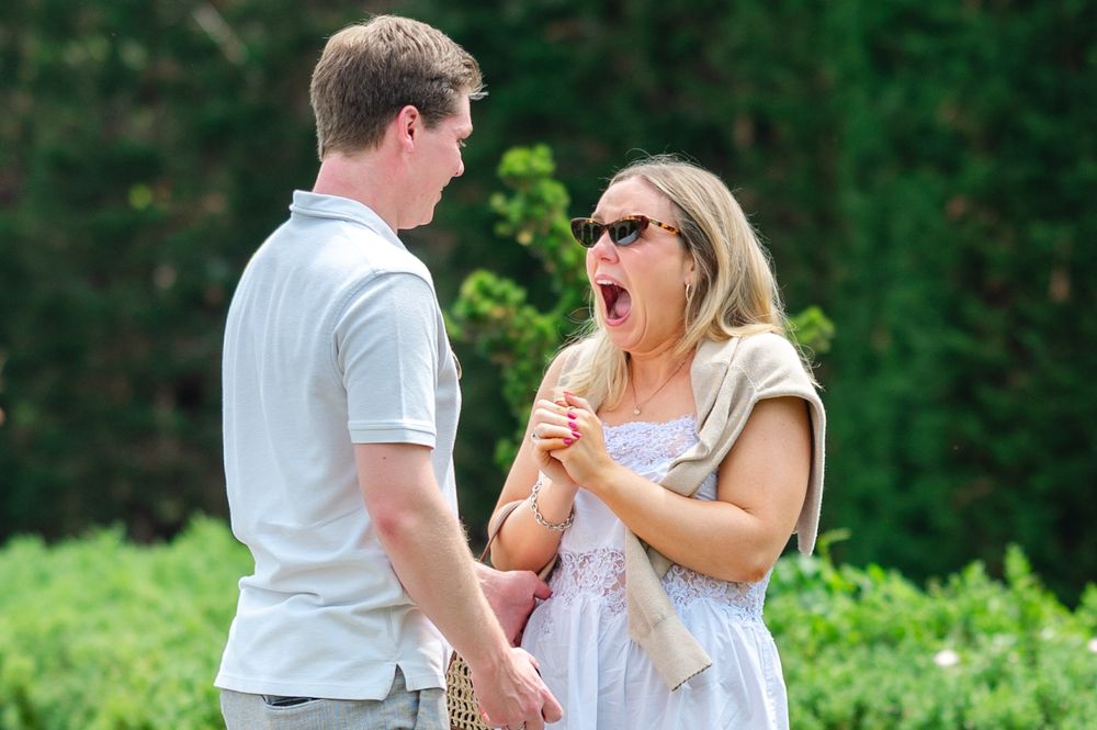 Surprised woman with sunglasses laughing and holding hands with a man outdoors in a lush green garden setting.