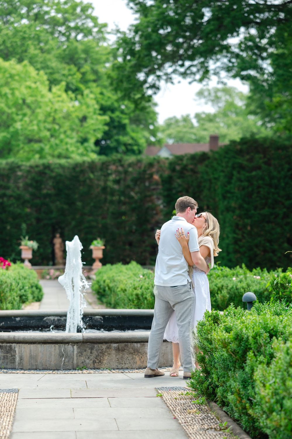 Couple sharing a romantic kiss near a garden fountain surrounded by lush green hedges.