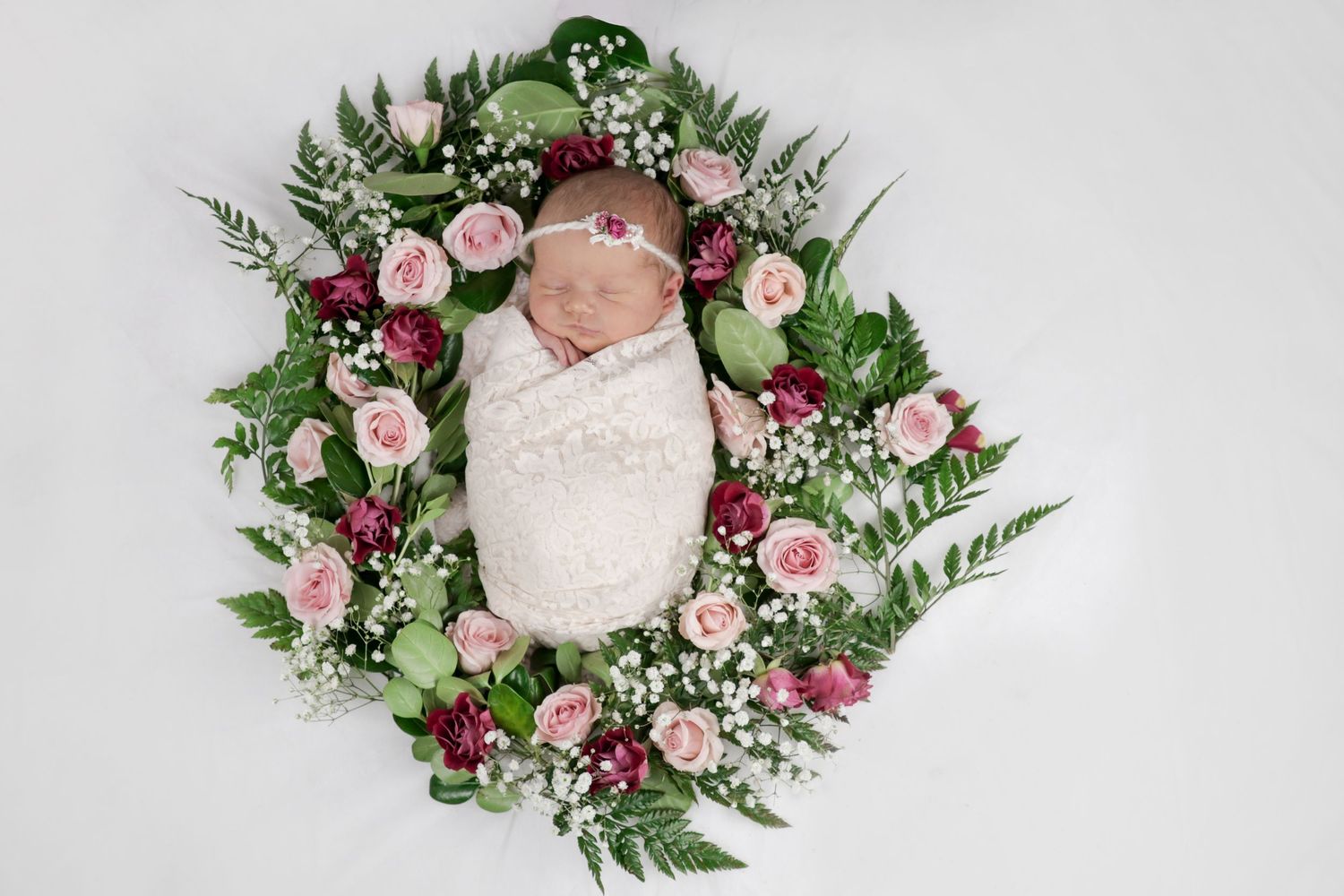 Newborn baby wrapped in white swaddle, nestled in a floral wreath of pink roses and greenery.