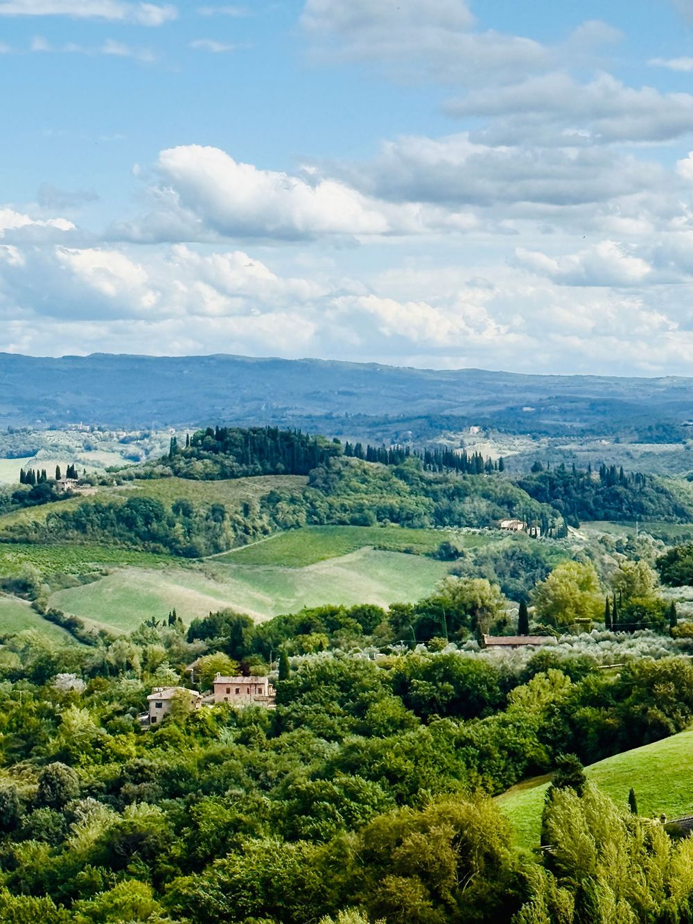 Panoramic aerial view of lush green rolling Tuscan hills with farmhouses under a cloudy blue sky.