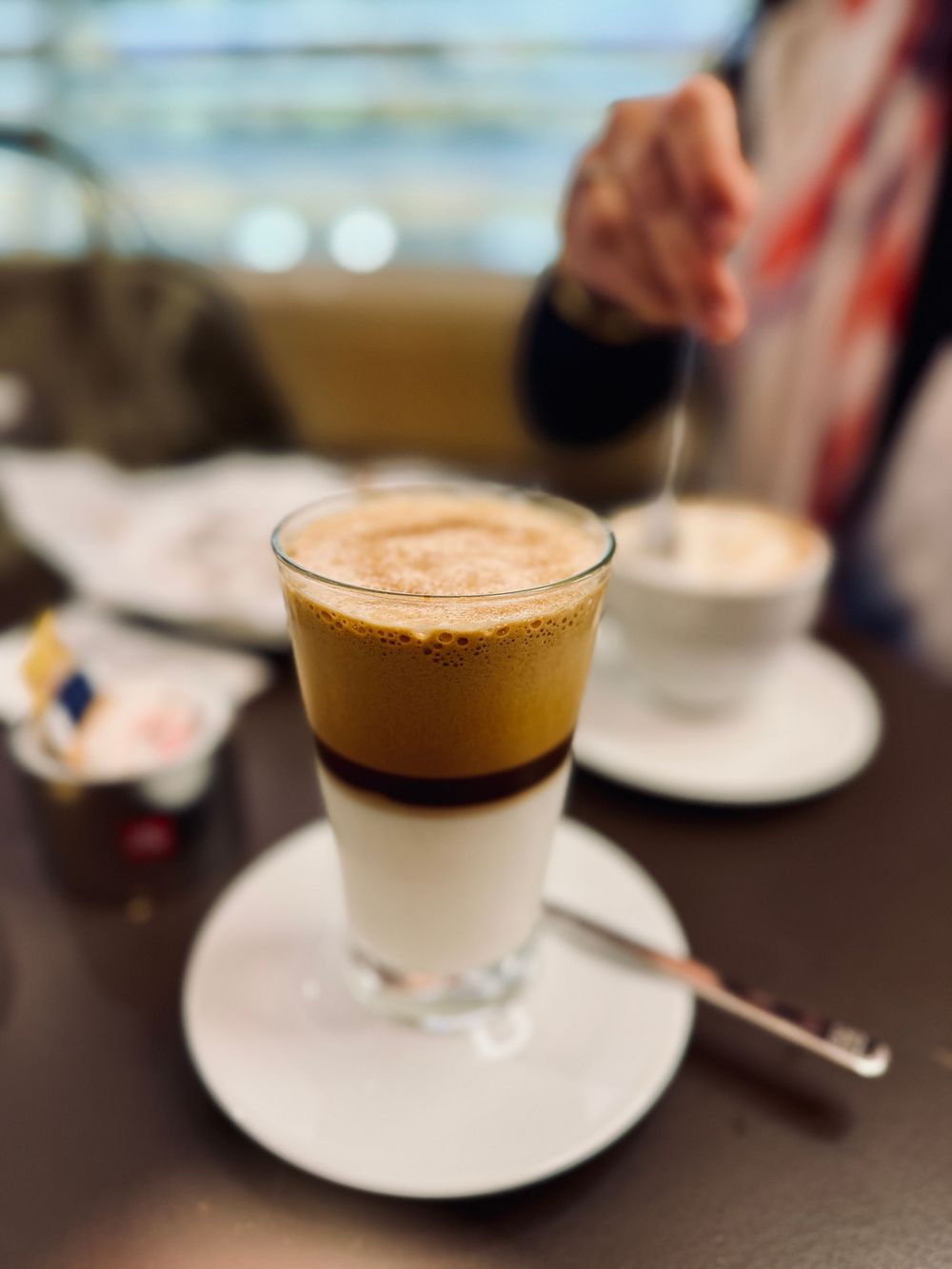 Layered espresso cortado coffee drink in a glass on a white saucer at a cafe table.
