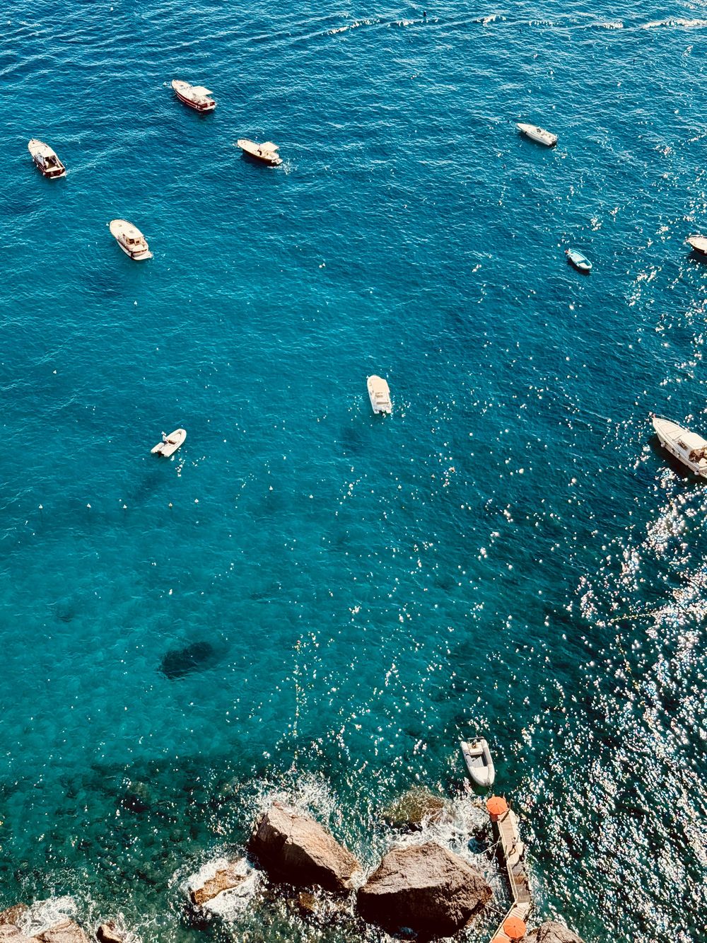 Aerial top-down view of small boats floating over vivid turquoise water near rocky Amalfi Coast shore.