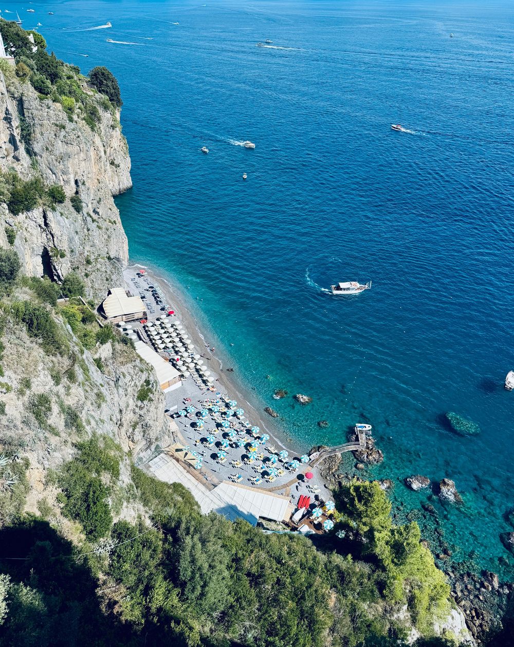 Aerial view of a small beach club with colorful umbrellas tucked beneath dramatic Amalfi Coast cliffs.