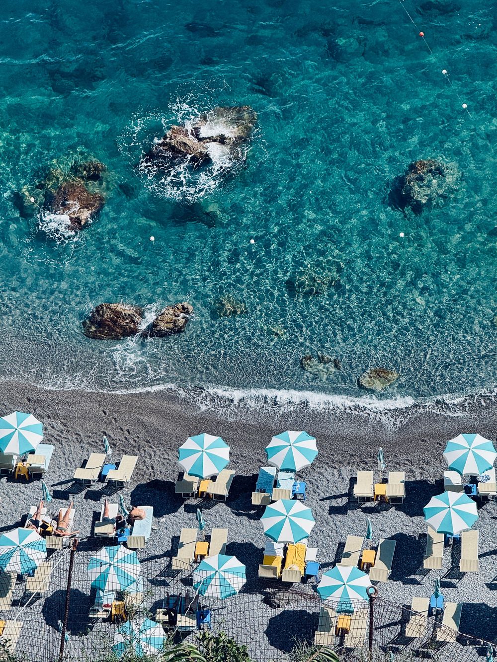 Top-down aerial view of blue and white beach umbrellas and lounge chairs along a rocky Mediterranean shore.