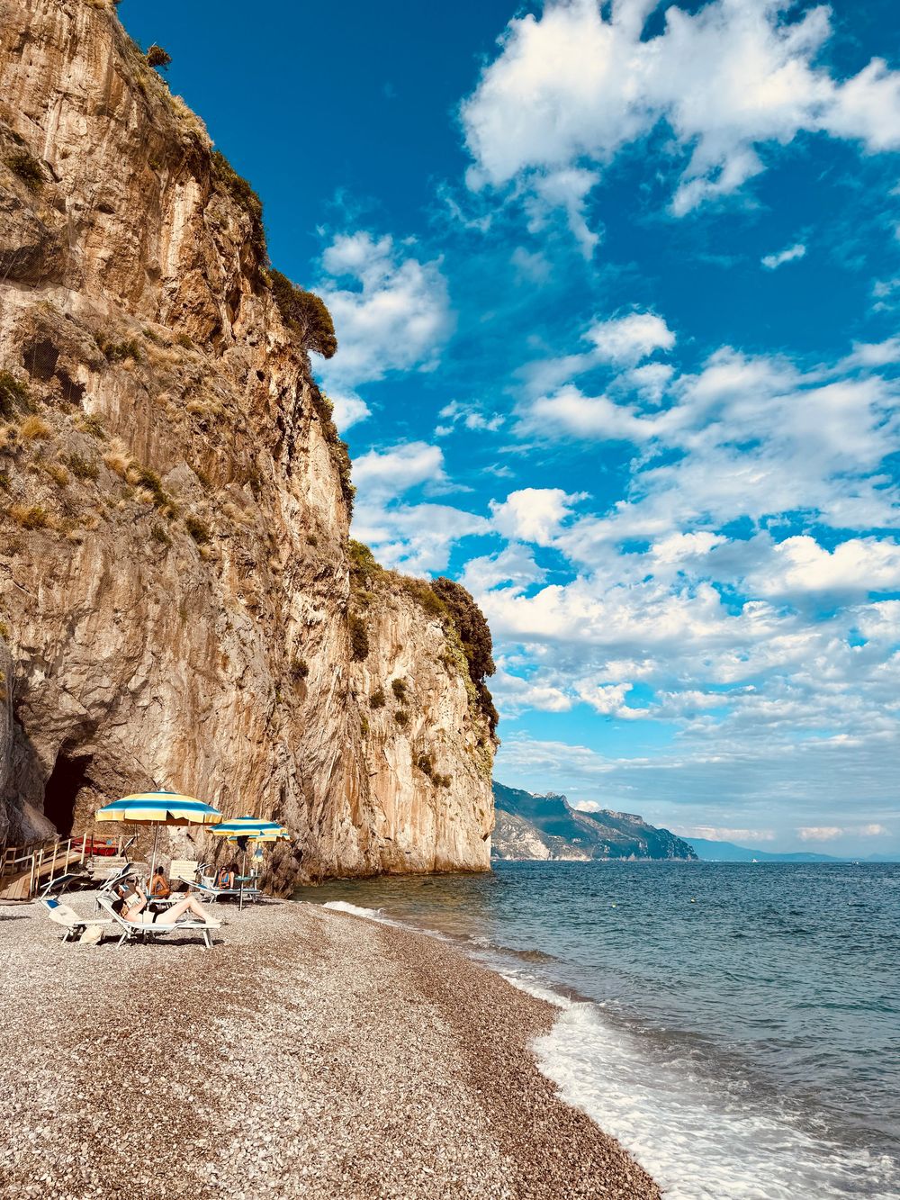 Rocky cliffside beach on the Amalfi Coast with colorful umbrellas, pebble shore, and blue Mediterranean sea.