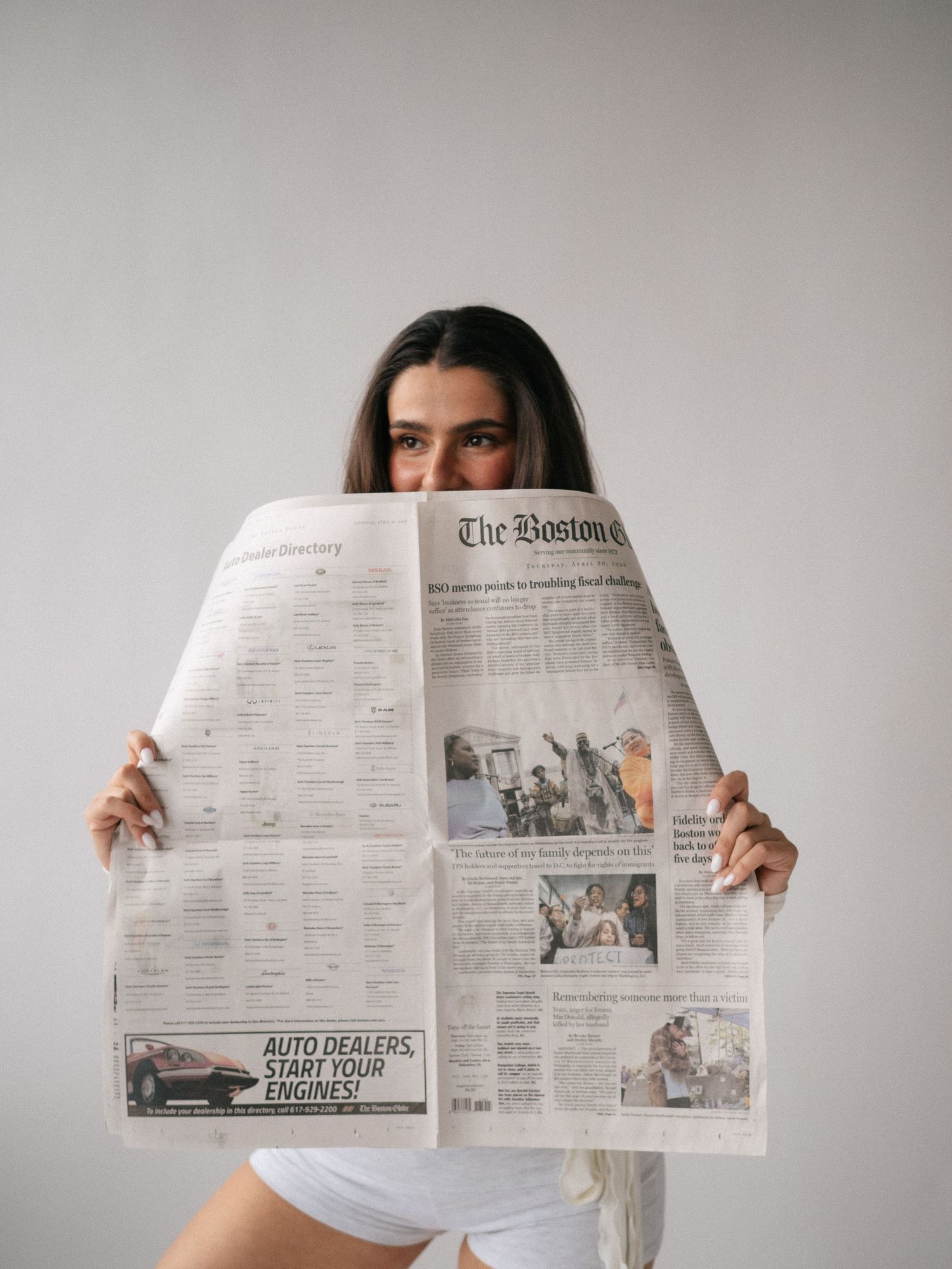 Woman holding The Boston Globe newspaper open, covering her face, wearing white outfit against light gray background.