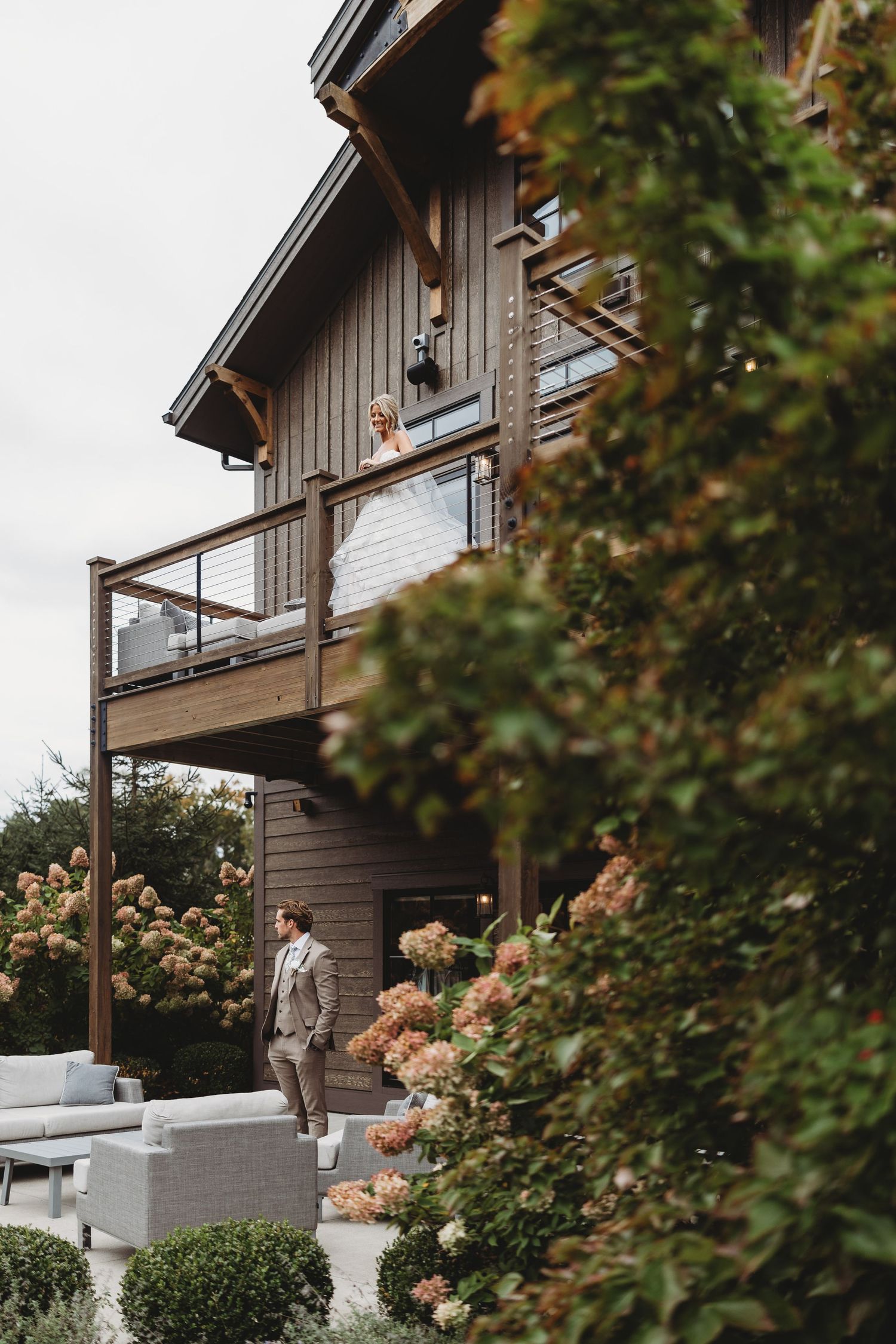A groom in a gray suit stands on a patio while the bride looks down from a wooden balcony surrounded by blooming hydrangeas.