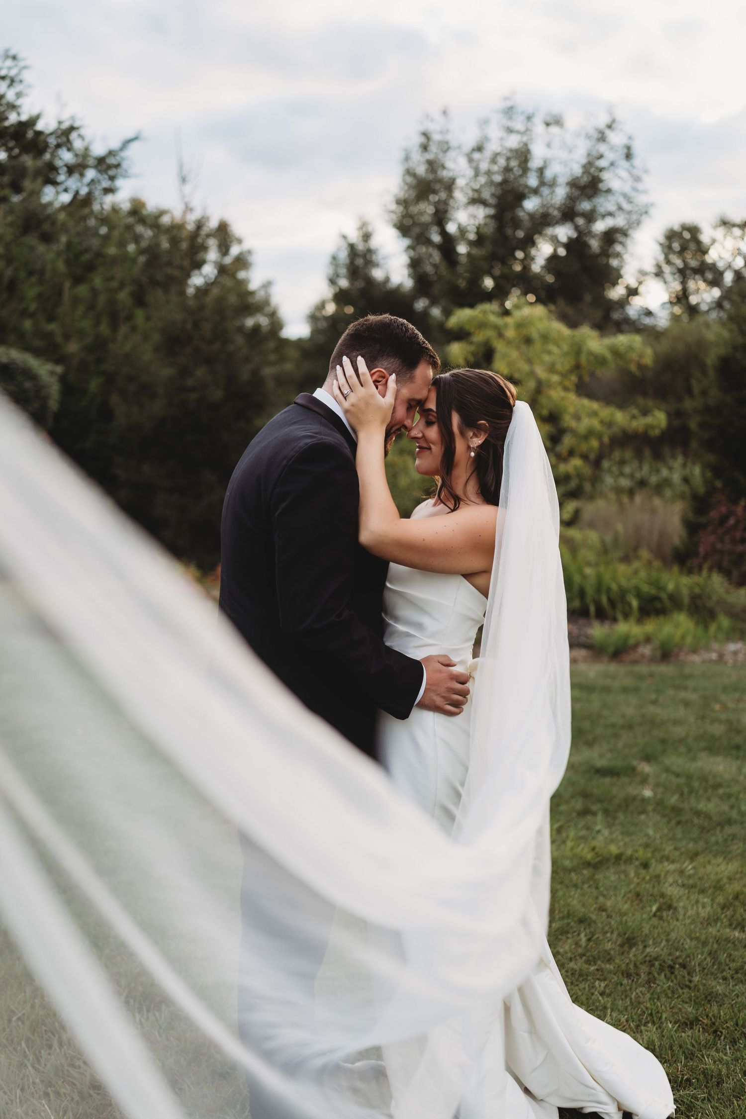 A bride and groom share an intimate moment outdoors, her veil flowing in the wind, surrounded by lush greenery.