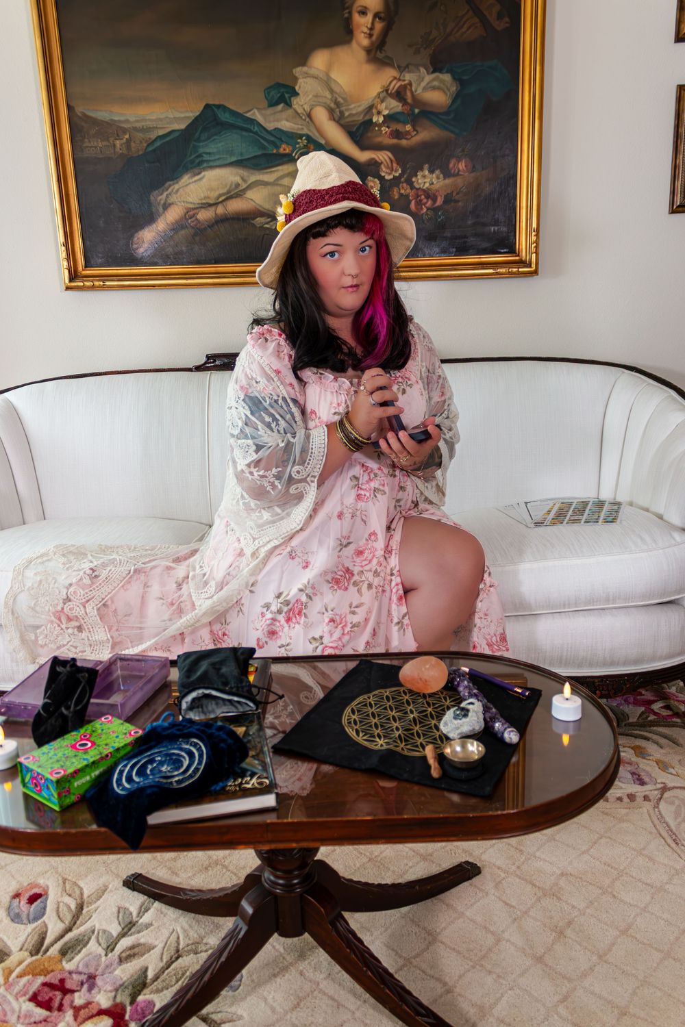 Woman in floral robe and hat sits on white sofa with tarot cards and crystals on wooden table.