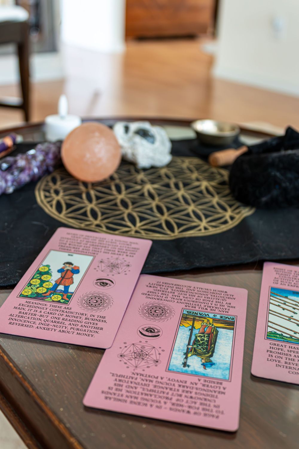 Pink tarot cards spread on wooden table with crystals, singing bowl, and flower of life mat display.