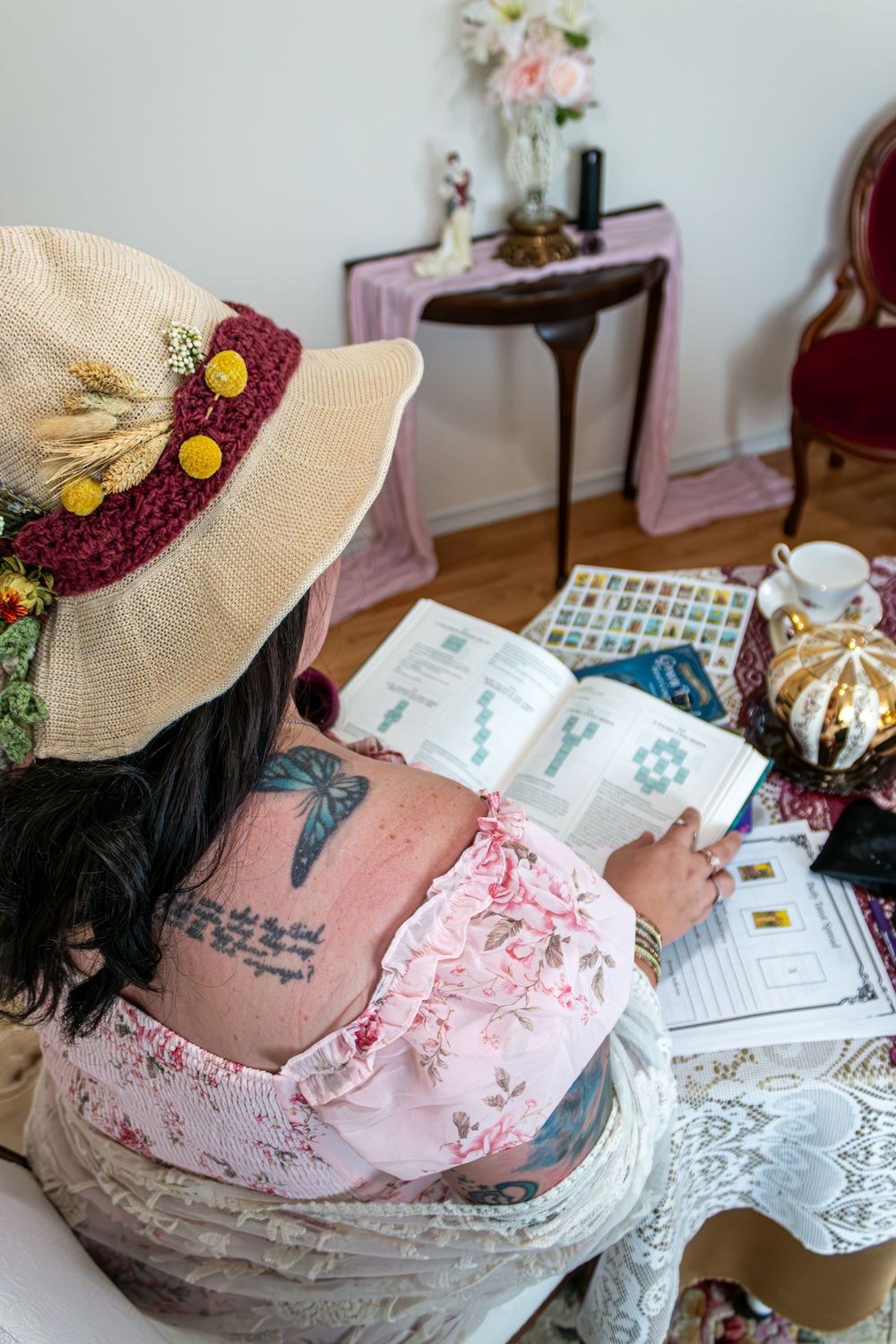 Woman with decorative hat and butterfly tattoo reading a book surrounded by colorful swatches and teacup.