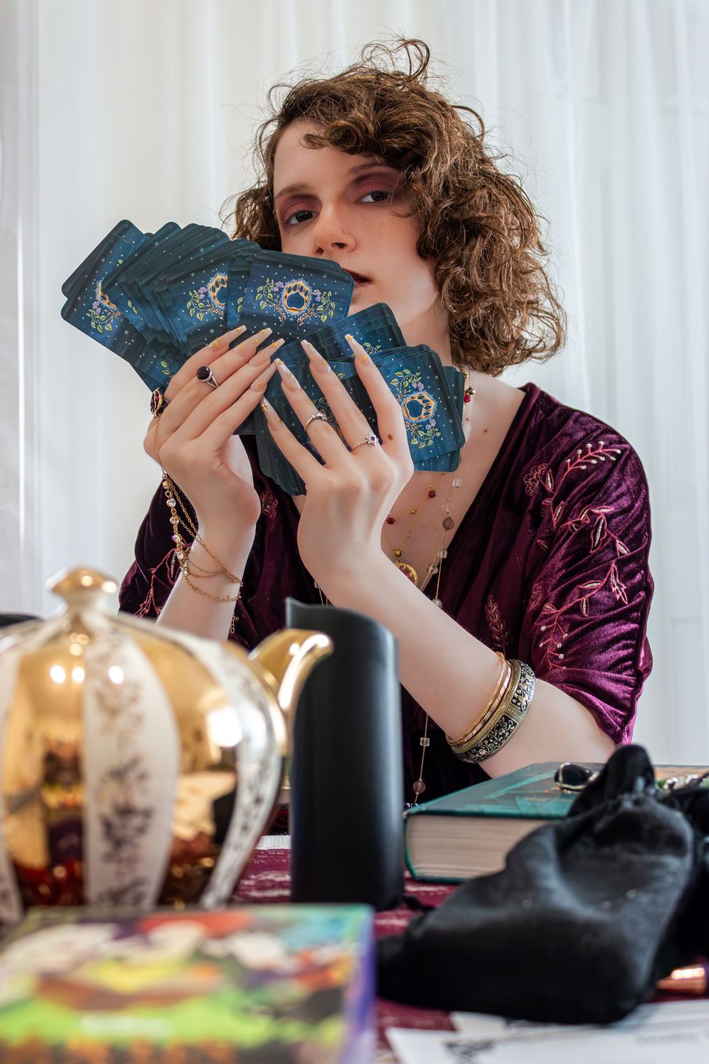 A tarot card reader in purple velvet holds a fan of blue tarot cards at a mystical table with candles and a teapot.