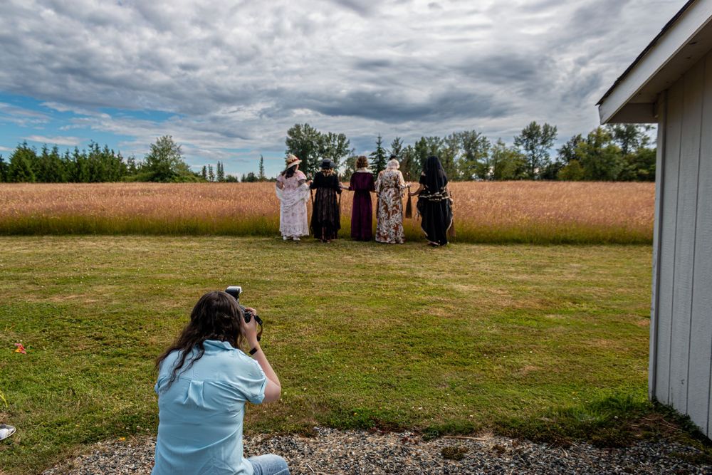 Photographer crouching to capture a group in formal attire posing outdoors near a golden field and white barn.