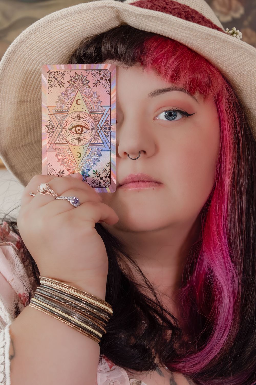 Woman with pink and black hair holding a holographic tarot card over her eye wearing a straw hat.
