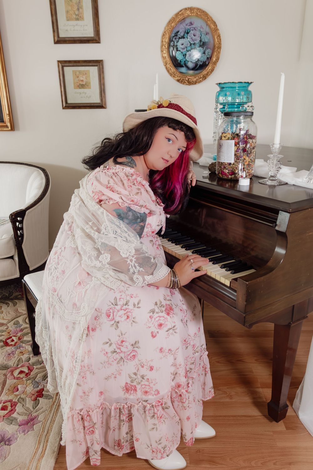 Woman in floral dress and hat playing an upright piano in a vintage-styled living room.