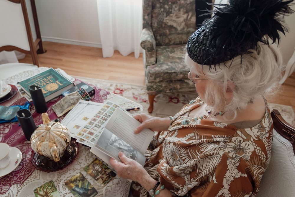 Elderly woman in vintage attire and black fascinator hat examining a stamp collection album at an antique-styled table.