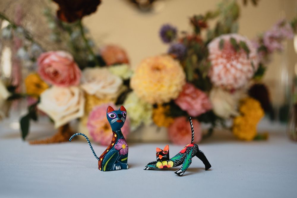 Two colorful Mexican alebrije cat figurines displayed on a table with vibrant floral arrangement in the background.