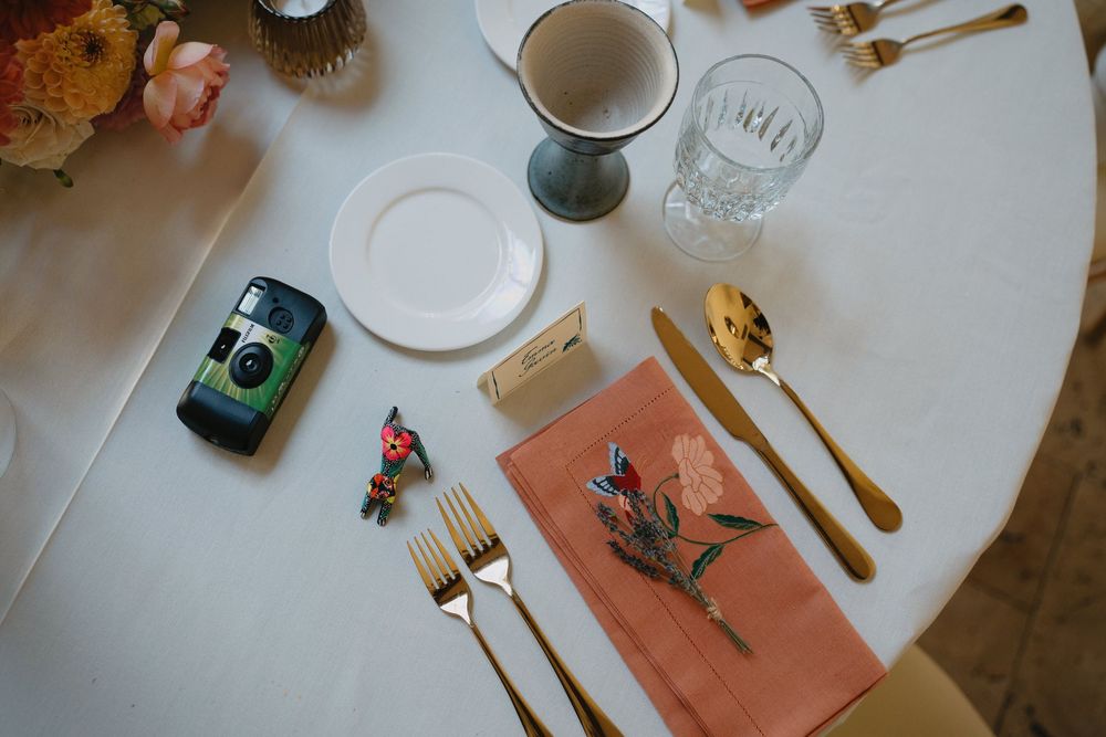 Elegant wedding table setting with gold cutlery, pink floral napkin, disposable camera, and glassware on white tablecloth.