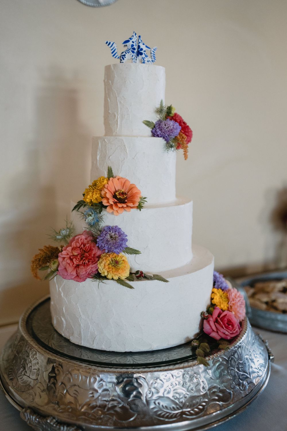 Four-tier white wedding cake adorned with colorful flowers and blue origami topper on silver stand.