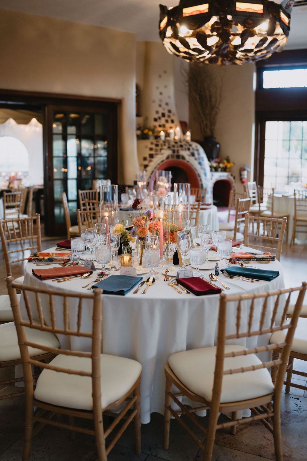 Elegant wedding reception table with white linens, colorful floral centerpiece, gold chiavari chairs, and a rustic fireplace backdrop.