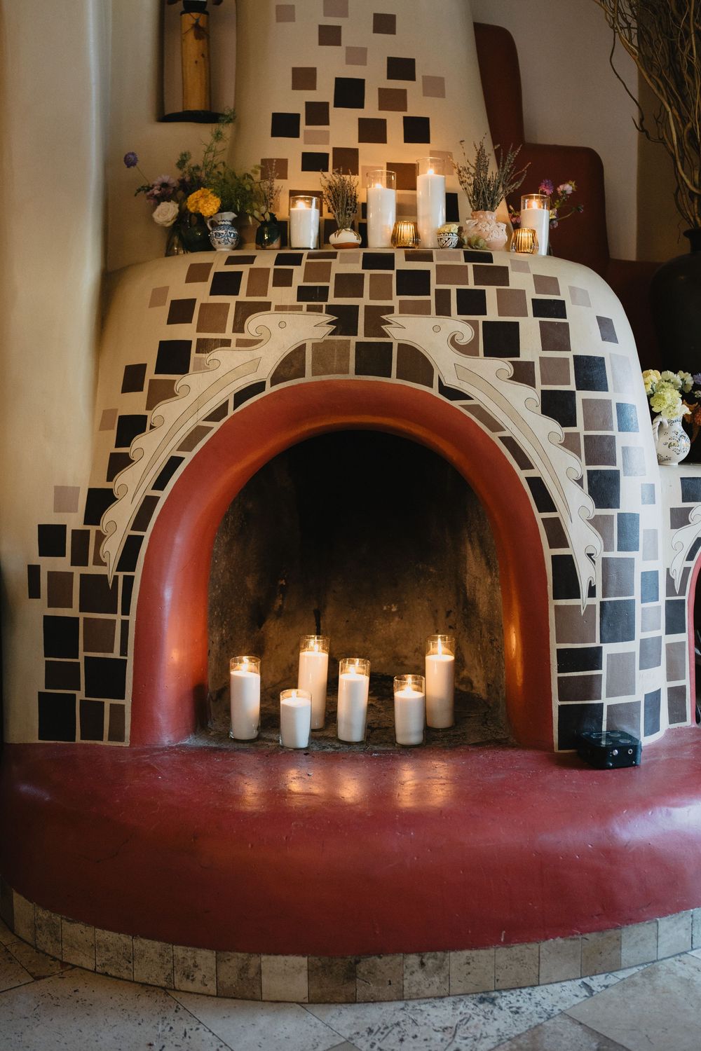Decorative tiled fireplace with white pillar candles inside and on the mantel, featuring a red arch and checkered mosaic tiles.