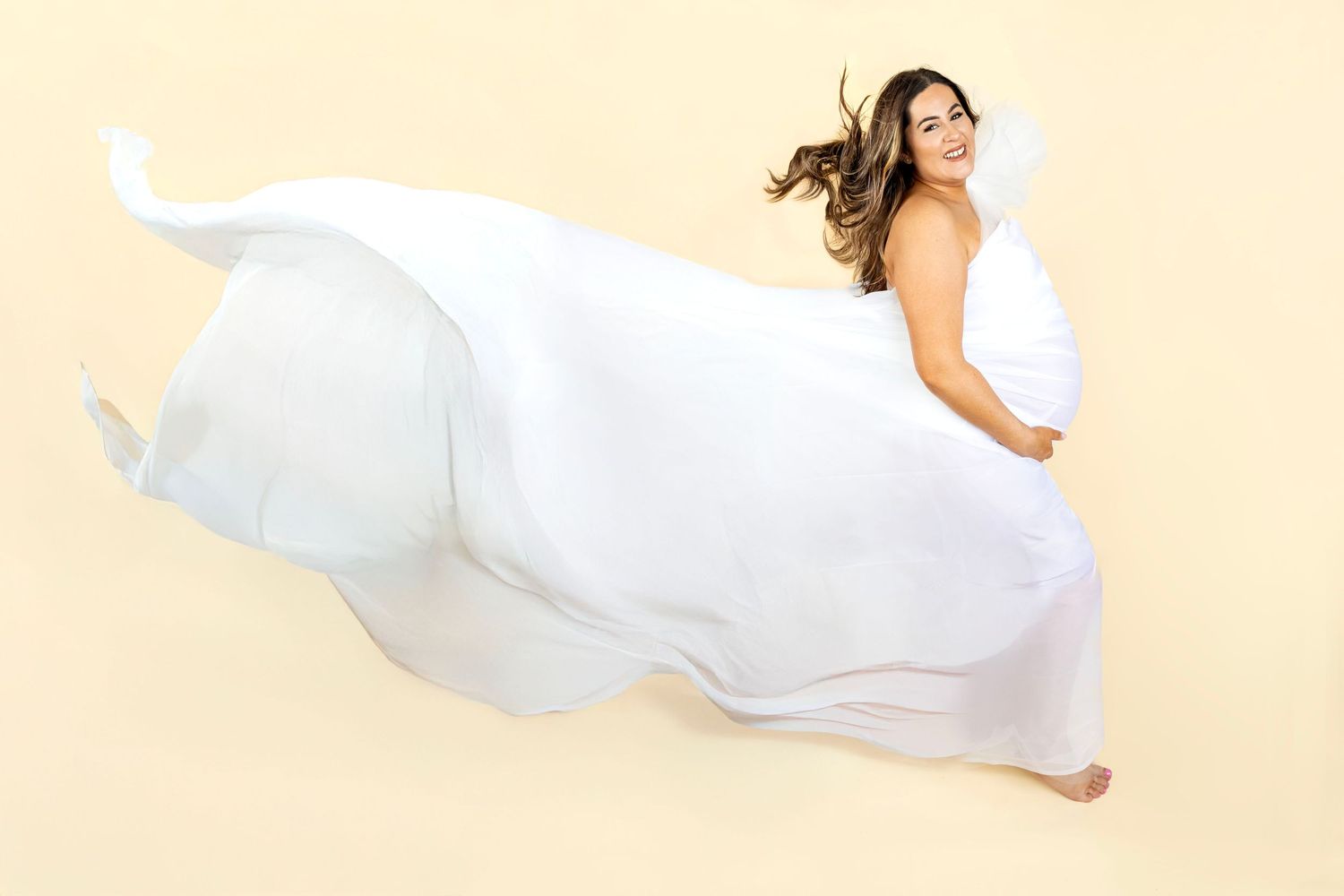 Pregnant woman in flowing white dress spinning gracefully against a soft beige background.R.A.M Photography, Stranraer