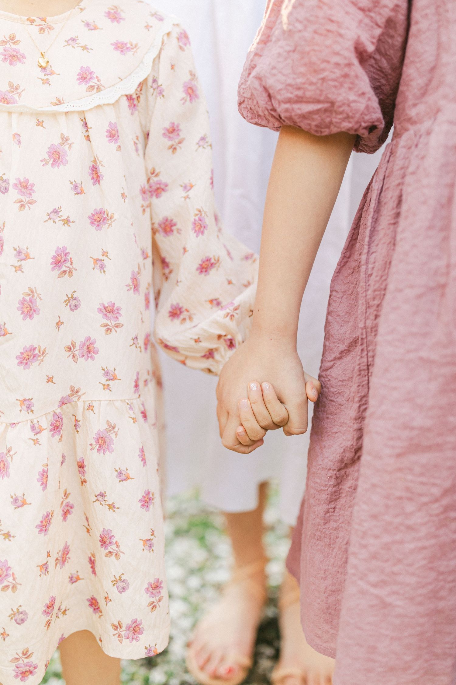 Two girls holding hands, one in a floral dress and one in a pink linen dress, standing barefoot outdoors.
